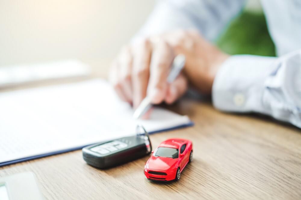 Car keys and a red toy car on a table, with man signing a car financing business loan in the background